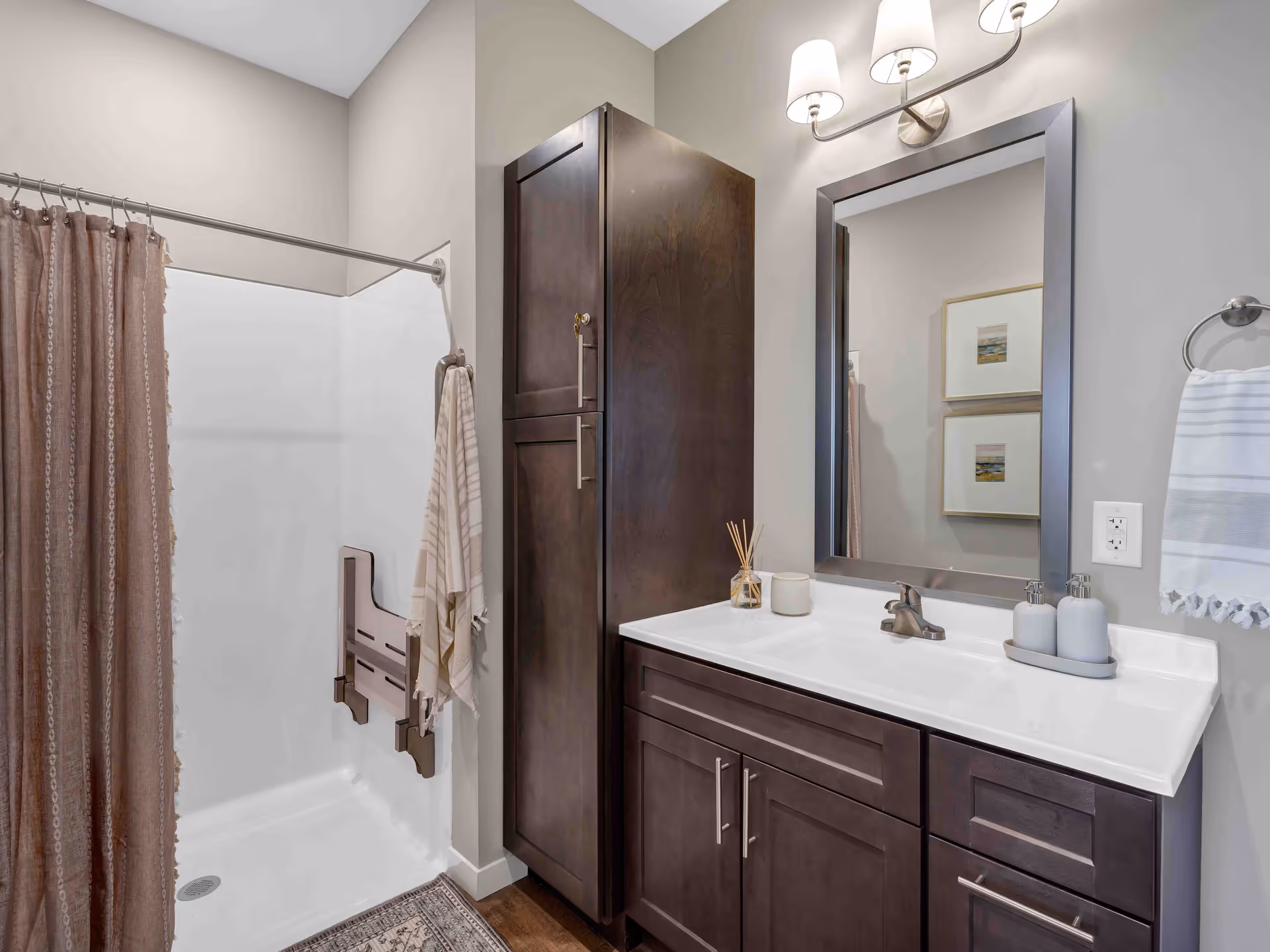 A modern bathroom featuring a walk-in shower with a brown shower curtain and a fold-down shower seat. There is a tall dark wood cabinet next to a white sink countertop with dark wood cabinetry below. Above the sink is a large rectangular mirror with a silver frame and a two-light wall sconce. A towel ring with a white towel is mounted on the wall next to the sink, and two framed pictures are reflected in the mirror.