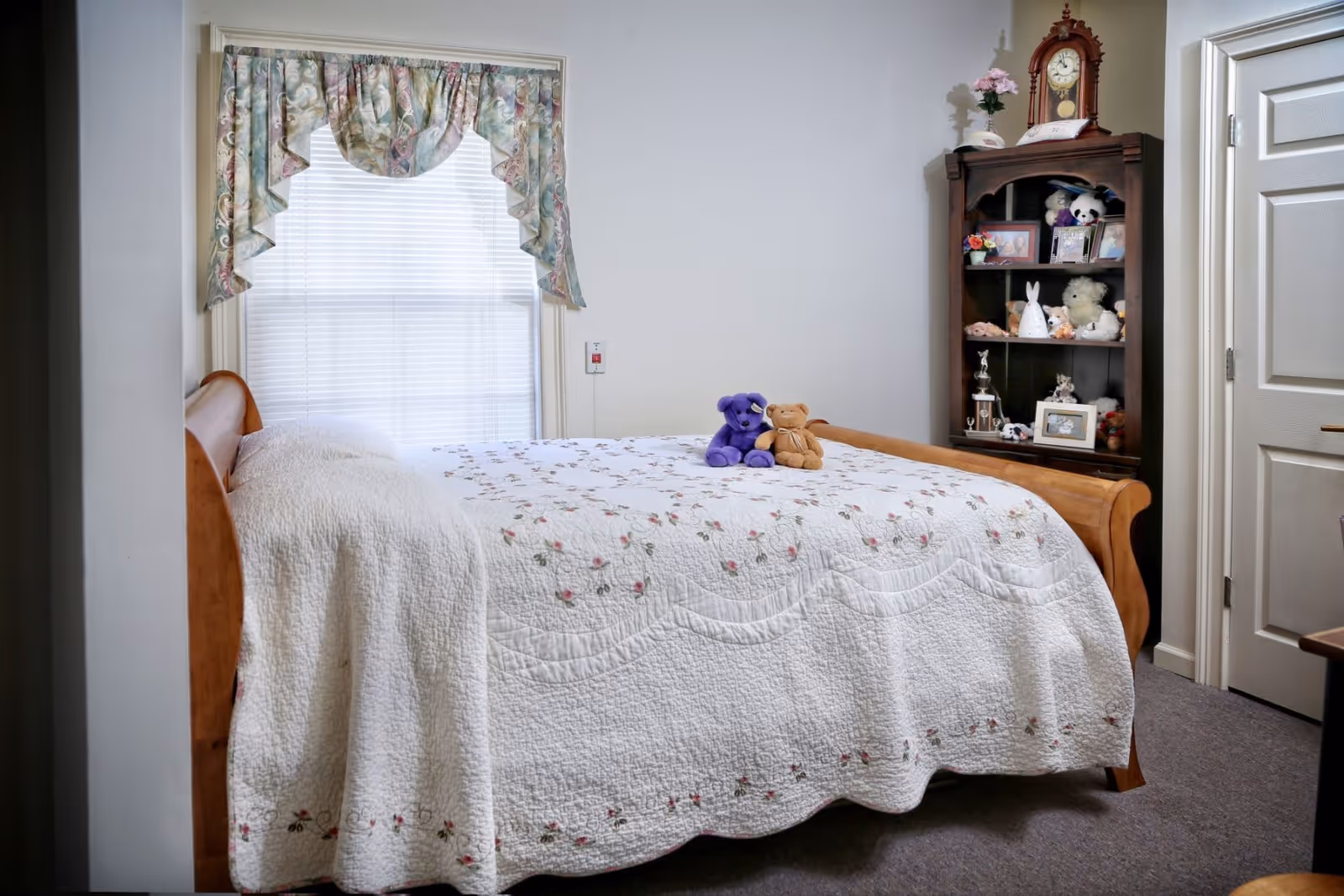 A cozy bedroom with a wooden bed covered by a white quilt with floral embroidery. Two stuffed bears, one purple and one brown, sit on the bed. A window with floral valance curtains is behind the bed. To the right, there is a wooden shelf filled with various stuffed animals, framed photos, and a clock on top. A closed white door is visible next to the shelf.