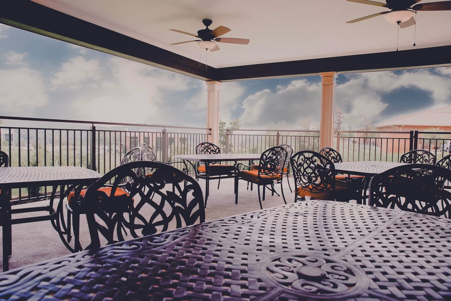 Covered outdoor patio with metal tables and chairs, ceiling fans, and a railing overlooking a landscaped view.