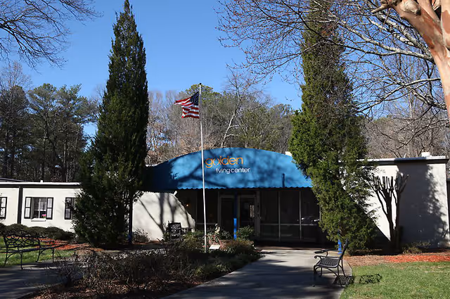 Exterior front view of a single-story building with a blue awning that reads 'golden living center'. There is an American flag on a flagpole in front of the entrance, surrounded by trees and benches along the walkway leading to the door.