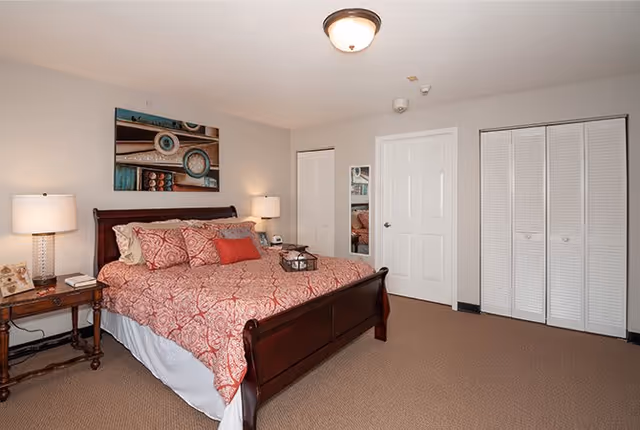 A bedroom with a large wooden bed featuring a red patterned bedspread and multiple pillows. There are two bedside tables with lamps on each side of the bed. The room has beige walls and carpeted floor. On the wall above the bed is a decorative painting. There are two white closet doors and a white door with a mirror mounted on it.