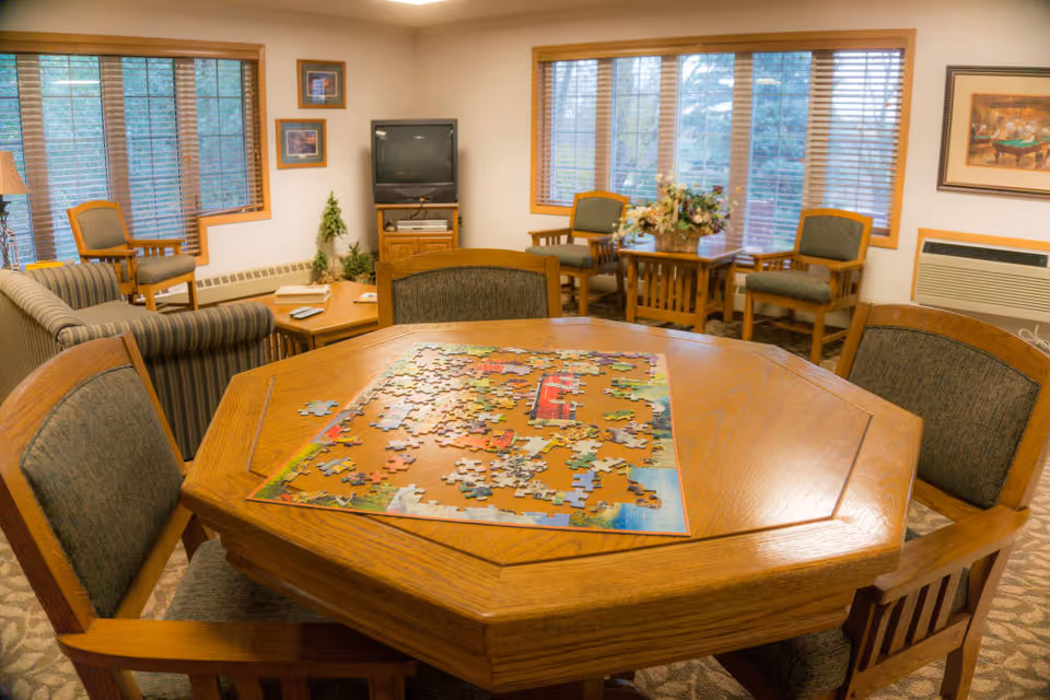 A cozy senior living common room with a wooden octagonal table in the foreground featuring a partially completed jigsaw puzzle. The room has several cushioned chairs around the table and additional seating including armchairs and a sofa near large windows with blinds. A television is placed on a wooden stand against the wall, and there is a small table with a floral arrangement next to two chairs by the window. The room is warmly lit and decorated with framed pictures on the walls.
