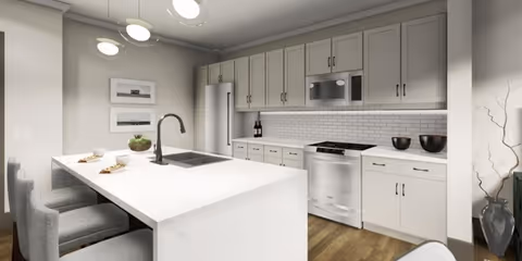 Modern kitchen with white cabinetry, a white countertop island with three gray upholstered chairs, stainless steel refrigerator, oven, and microwave. The kitchen features a white subway tile backsplash and wooden flooring. There are decorative items including two bottles of wine, a plant, and bowls on the countertop.