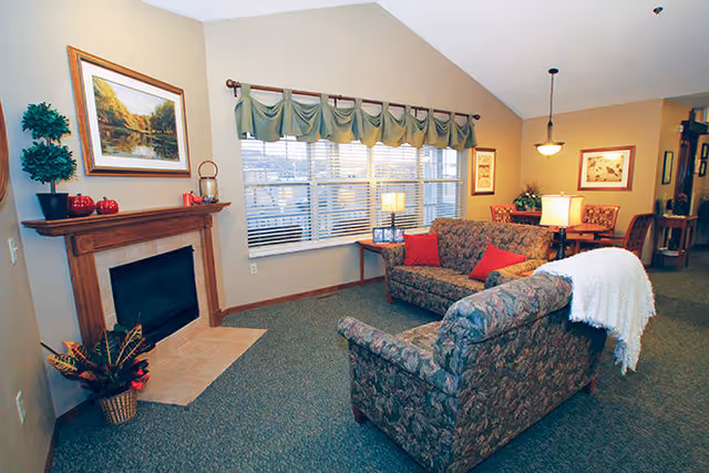 A cozy living room with two patterned sofas, one with a white throw blanket and red cushions. There is a wooden fireplace with decorative items and a framed landscape painting above it. A large window with green valances lets in natural light. The room also features a side table with a lamp and a dining area visible in the background.