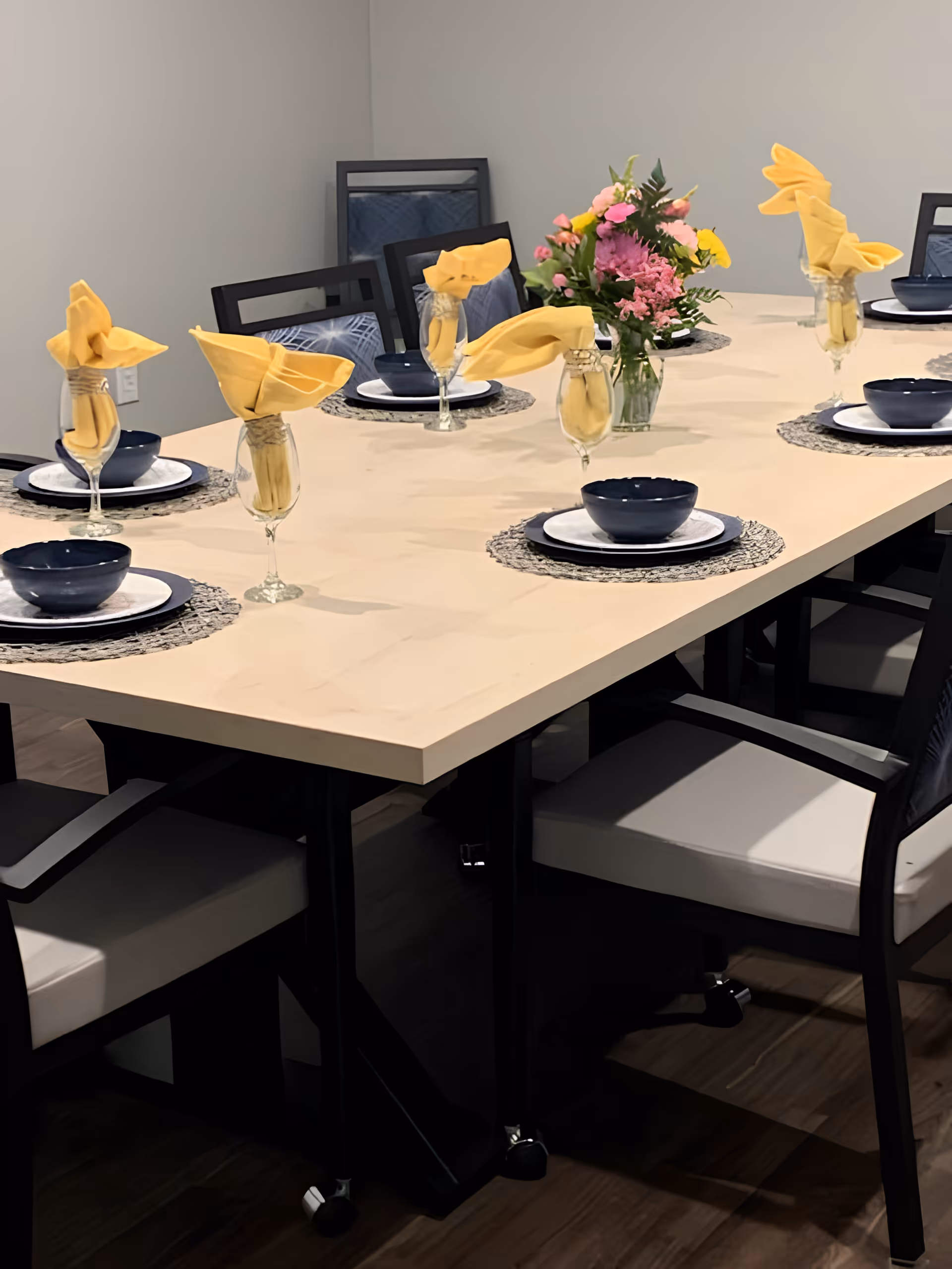 A long dining table set with plates, navy bowls, wine glasses holding folded yellow napkins and a floral centerpiece.