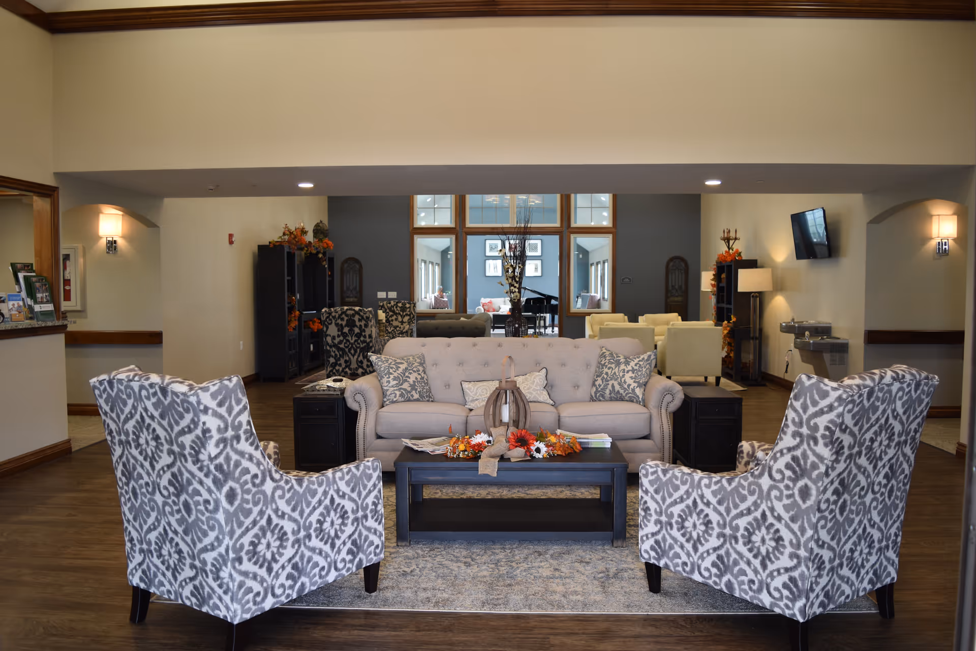 A cozy senior living facility common area with a beige sofa and two patterned armchairs arranged around a black coffee table decorated with autumn-themed items. The room has wooden flooring, soft lighting from wall sconces, and a large windowed wall in the background revealing another seating area with cream-colored chairs and a piano.