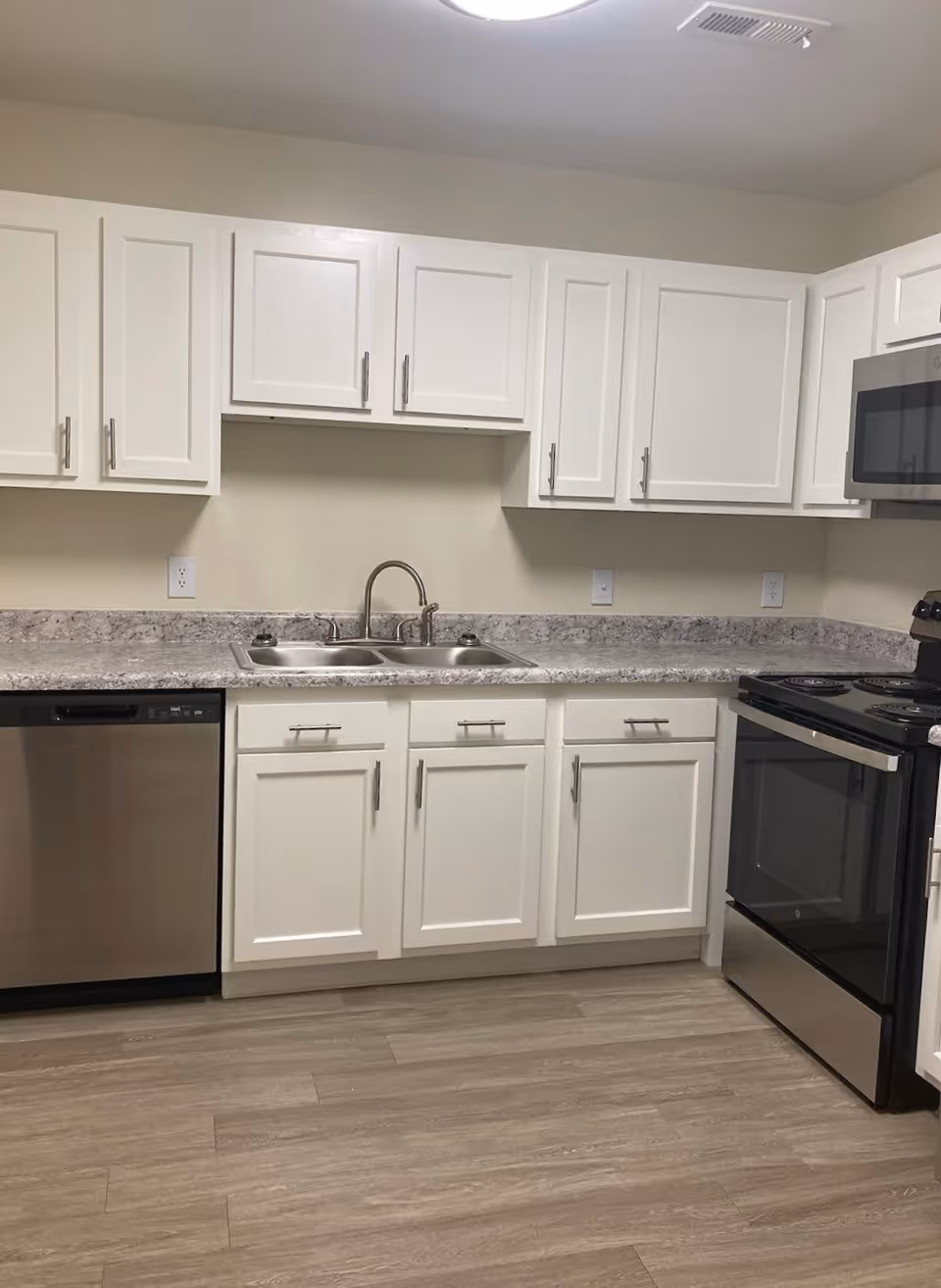 A modern kitchen with white cabinets, a double stainless steel sink, a stainless steel dishwasher, a black and stainless steel electric stove, and a microwave. The countertop is gray and white granite, and the floor is light wood.
