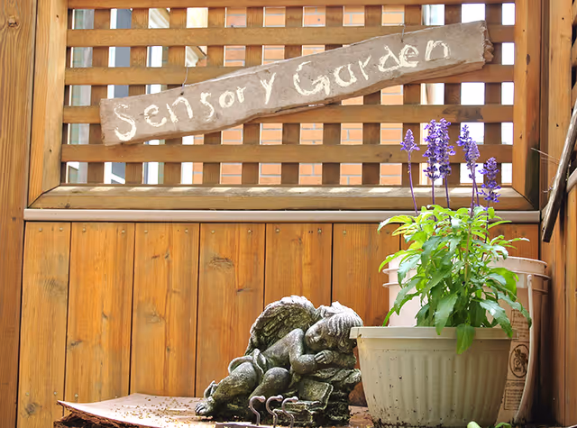 A small wooden sign reading 'Sensory Garden' hangs on a lattice wooden fence. Below the sign, there is a small statue of a sleeping angel lying on its side next to a white pot with green plants and purple flowers.