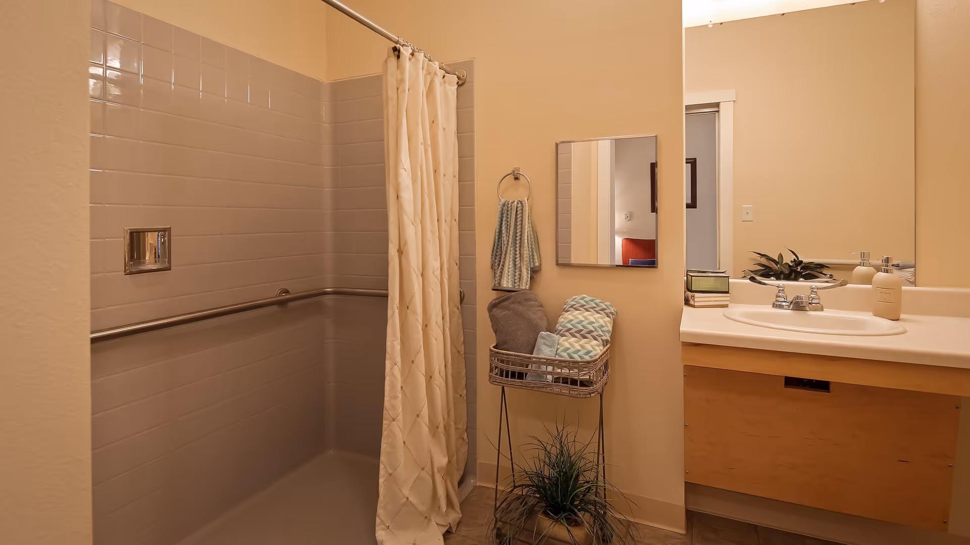A bathroom with beige walls featuring a tiled shower area with a white shower curtain and a metal grab bar. Next to the shower is a small mirror above a towel ring holding two towels. Below the mirror is a metal basket stand with folded towels and a small plant on the floor. To the right is a sink with a large mirror above it, soap dispensers, and a small decorative plant.