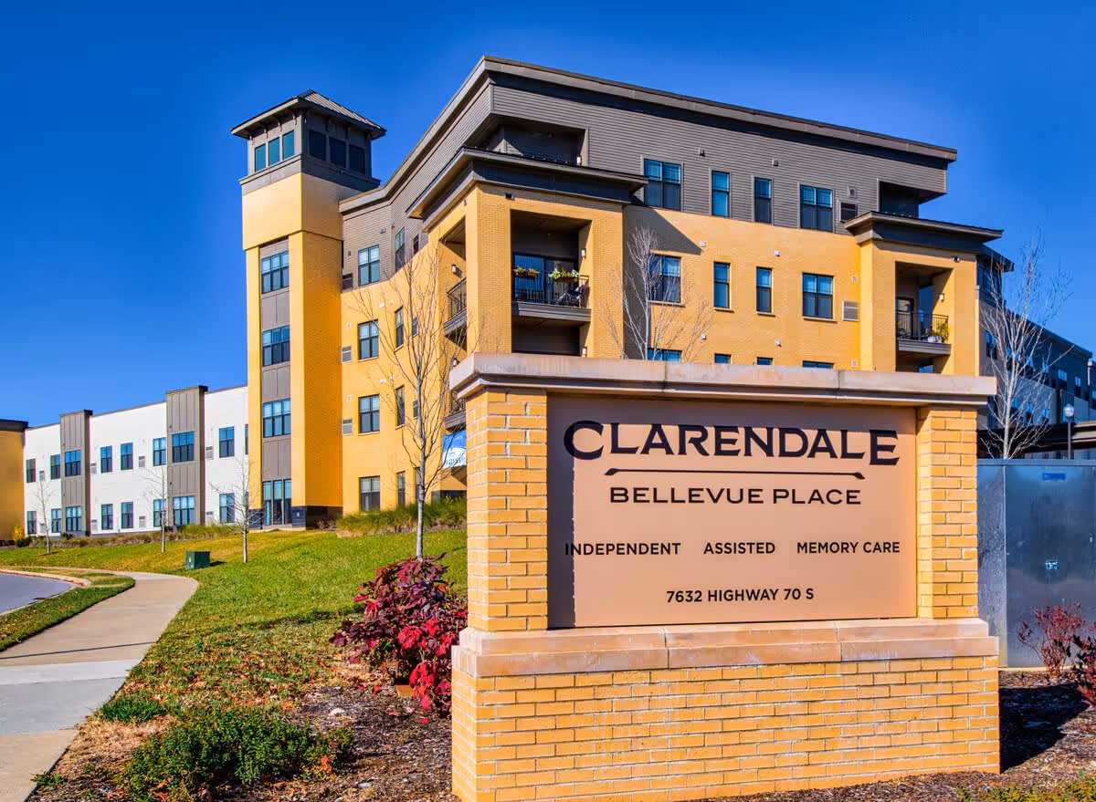 Exterior view of Clarendale at Bellevue Place, a multi-story senior living facility with yellow and white walls, balconies, and a tower-like structure. In the foreground, there is a brick sign displaying the facility's name, services offered (Independent, Assisted, Memory Care), and address (7632 Highway 70 S). The sky is clear and blue.