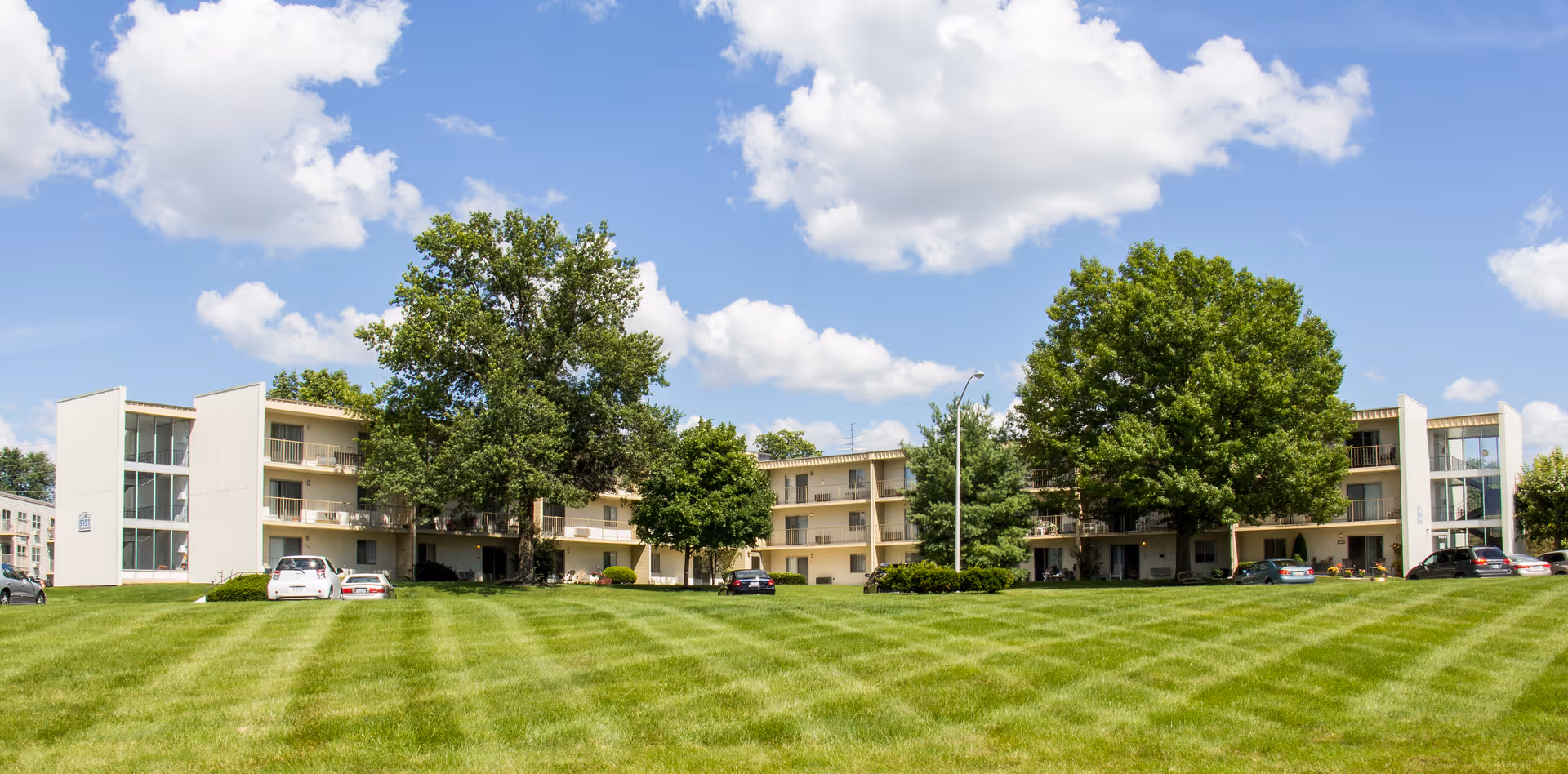 Exterior view of a three-story senior living facility building with balconies, surrounded by large green trees and a well-maintained lawn under a partly cloudy blue sky. Several cars are parked near the building.