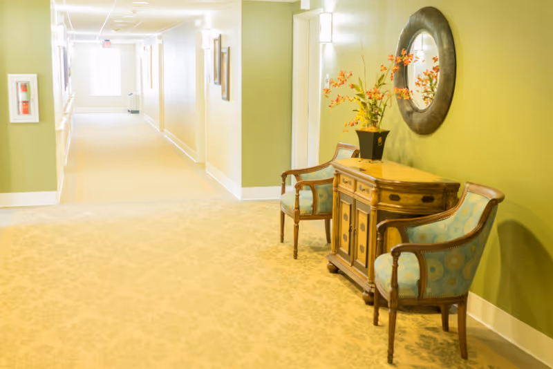 A well-lit hallway with light green walls and beige carpet. On the right side, there is a wooden console table with a vase of orange flowers and two upholstered chairs with wooden frames. Above the table is a round decorative mirror. The hallway extends into the distance with framed pictures on the walls and an exit sign at the far end.