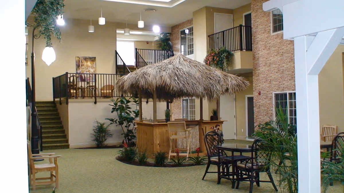 Indoor common area with a thatched-roof tiki bar, tables and chairs, plants, and upper-level balconies.