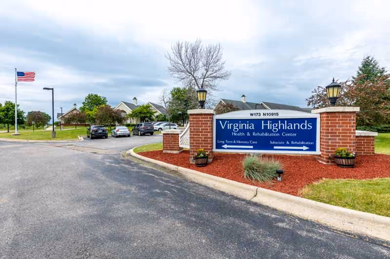 Entrance sign for Virginia Highlands Health & Rehabilitation Center mounted on brick pillars at the driveway entrance with buildings and an American flag in the background.