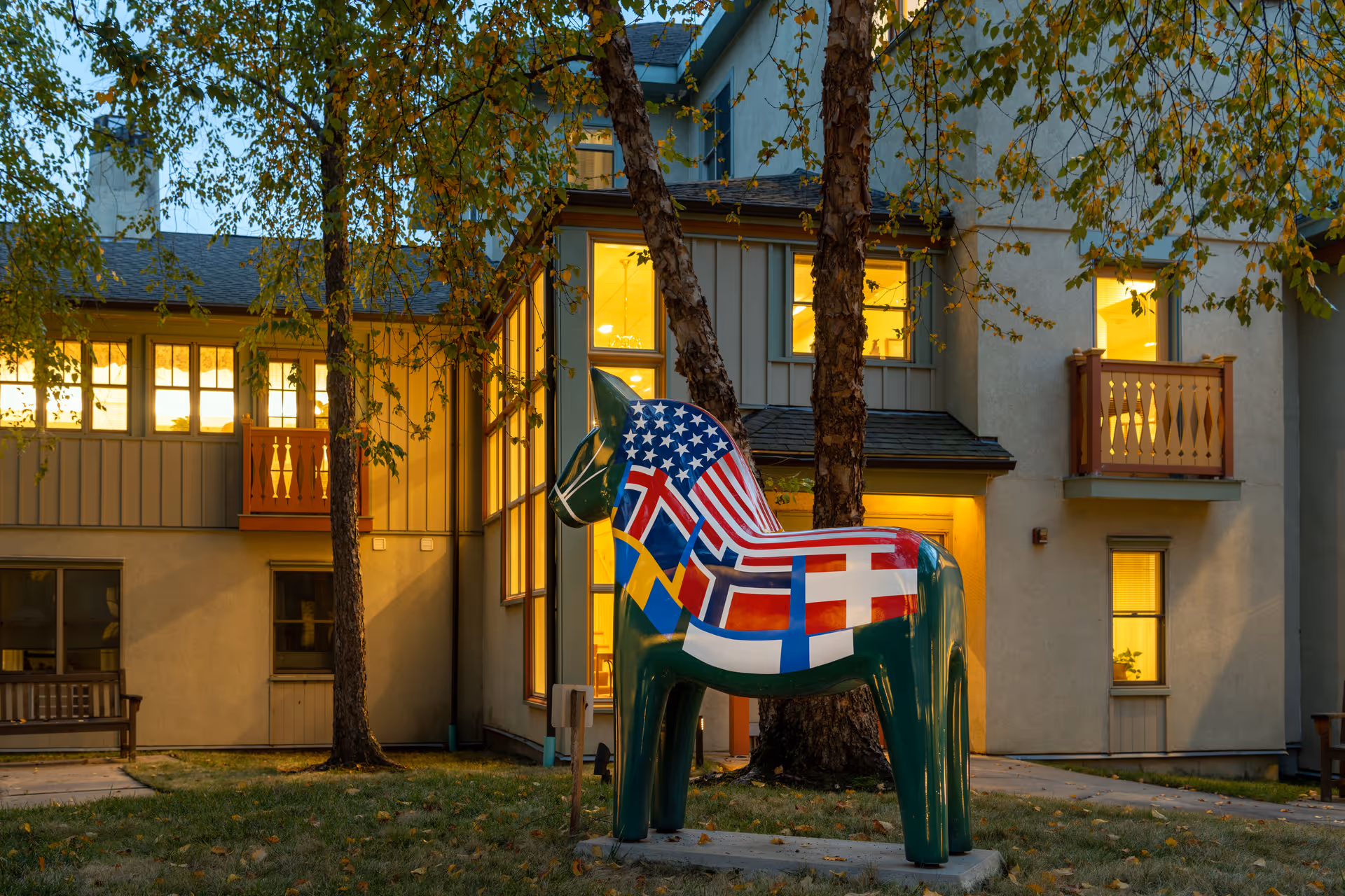 Colorful painted horse statue covered with international flags stands on the grass in front of a lit multi-story residential building at dusk.