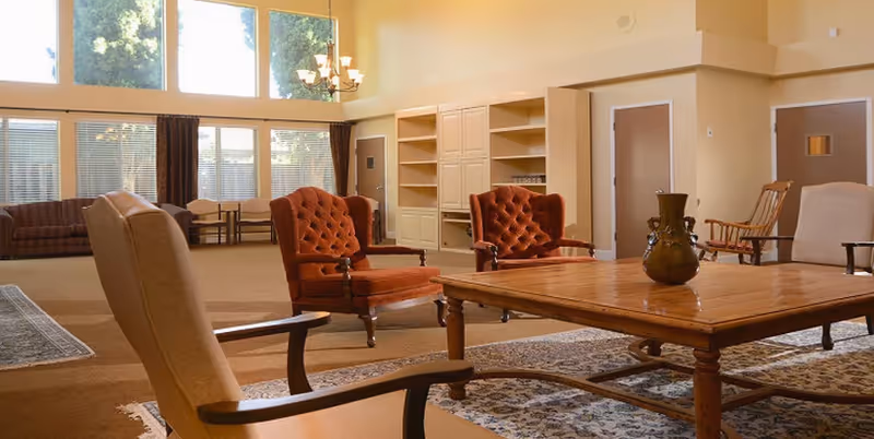 A spacious living room with large windows letting in natural light, featuring a wooden coffee table with a decorative vase, several upholstered armchairs in beige and rust colors, a rocking chair, and built-in shelves and cabinets along the wall.