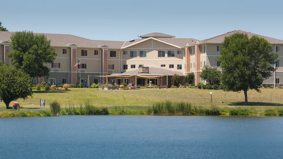 A senior living center fronts a lake, with manicured lawn, trees, and a seating area.