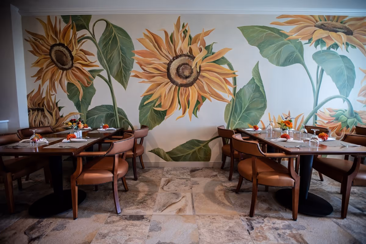 Dining area with two wooden tables and brown cushioned chairs set with placemats, napkins, and small floral centerpieces. The background features a large mural of sunflowers and green leaves painted on the wall.