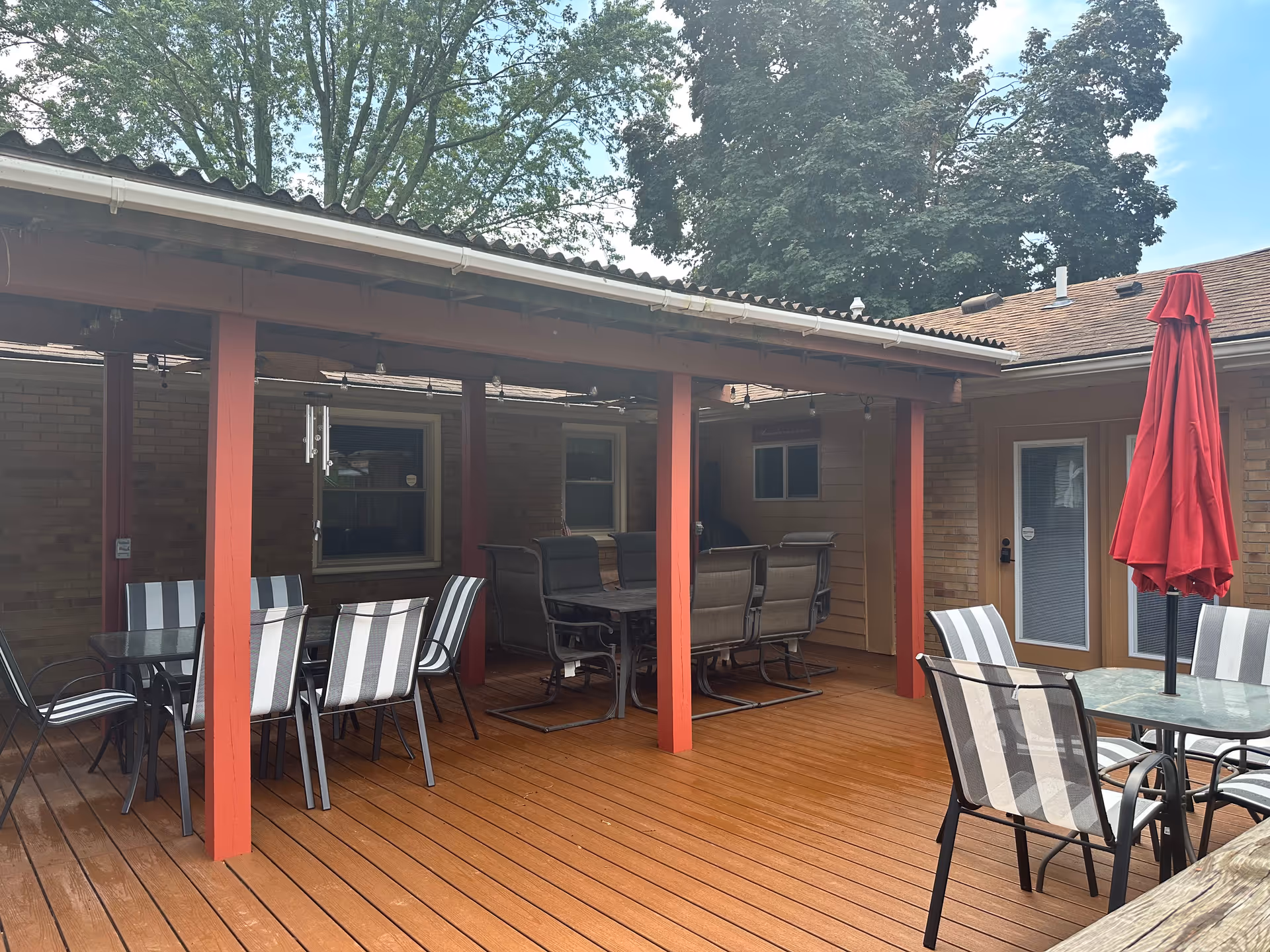 Outdoor patio area with wooden deck flooring, several tables with striped chairs, a red umbrella, and a covered seating area supported by red wooden posts. Trees and a partly cloudy sky are visible in the background.