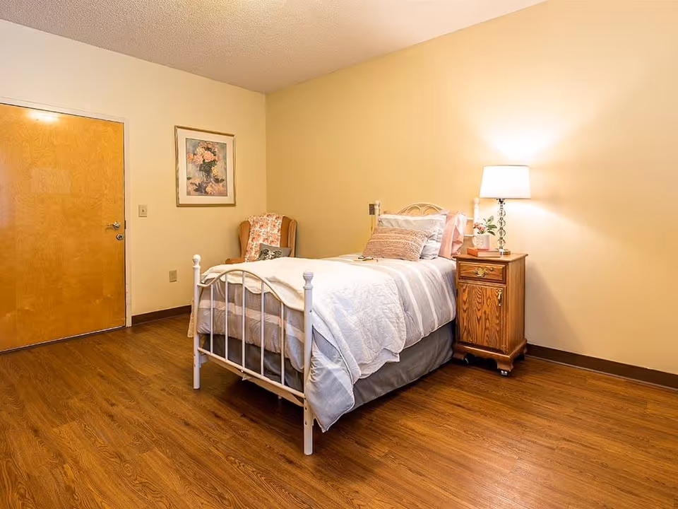 A simple bedroom with a single white metal bed frame, neatly made with white and gray bedding and multiple pillows. Next to the bed is a wooden nightstand with a lamp, a small plant, and a book. There is a wooden door on the left wall, a floral painting above a cushioned armchair with a floral blanket draped over it. The room has wooden flooring and beige walls.