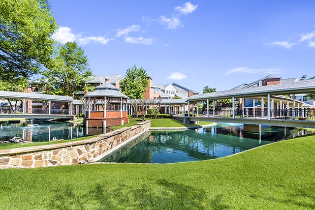 Outdoor view of Brookdale Club Hill featuring a landscaped pond with a stone retaining wall, a wooden gazebo, covered walkways, green grass, and residential buildings in the background under a blue sky with scattered clouds.