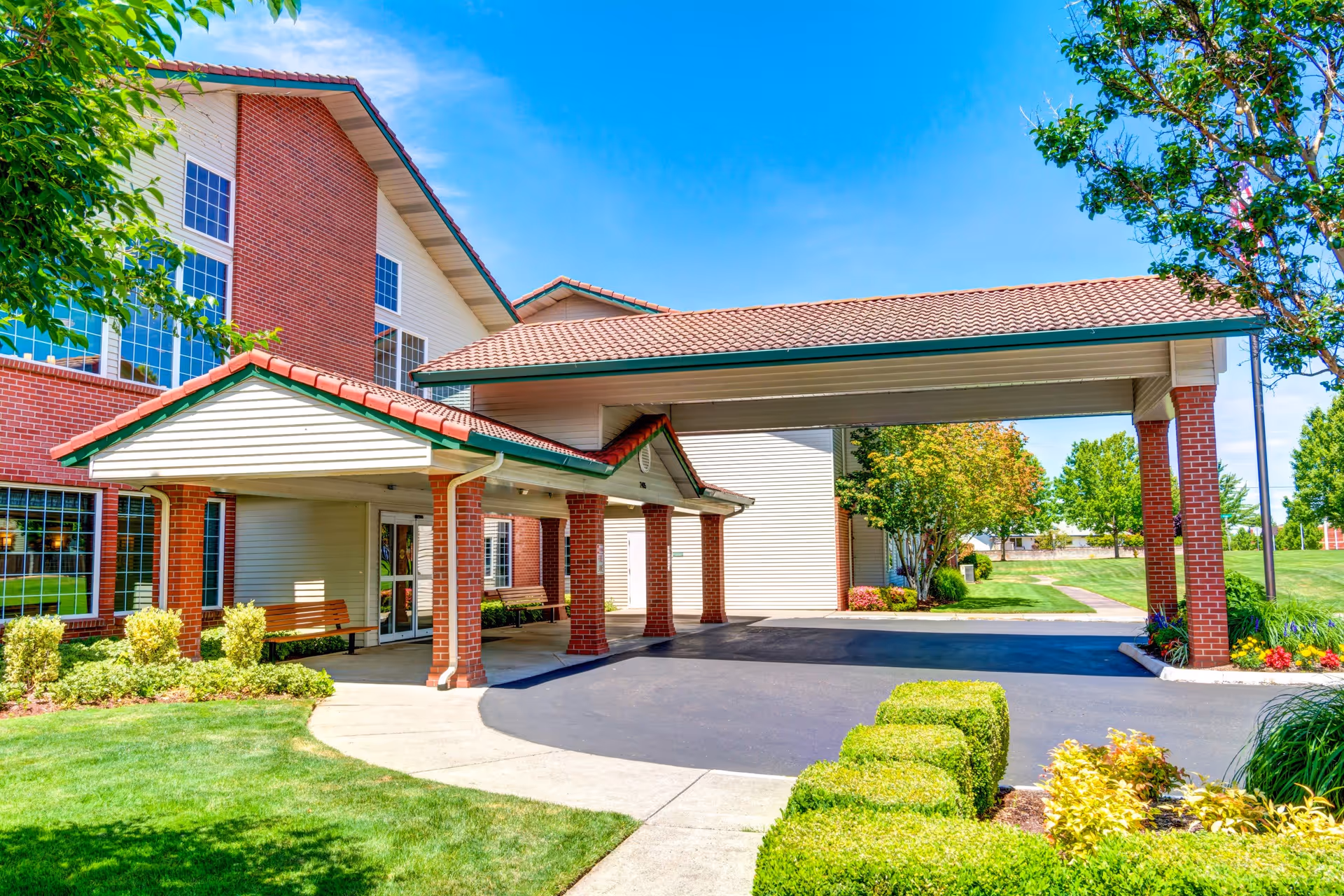 Exterior view of Rosewood Park facility entrance with a covered drop-off area supported by brick columns, surrounded by well-maintained landscaping including green grass, bushes, and trees under a clear blue sky.