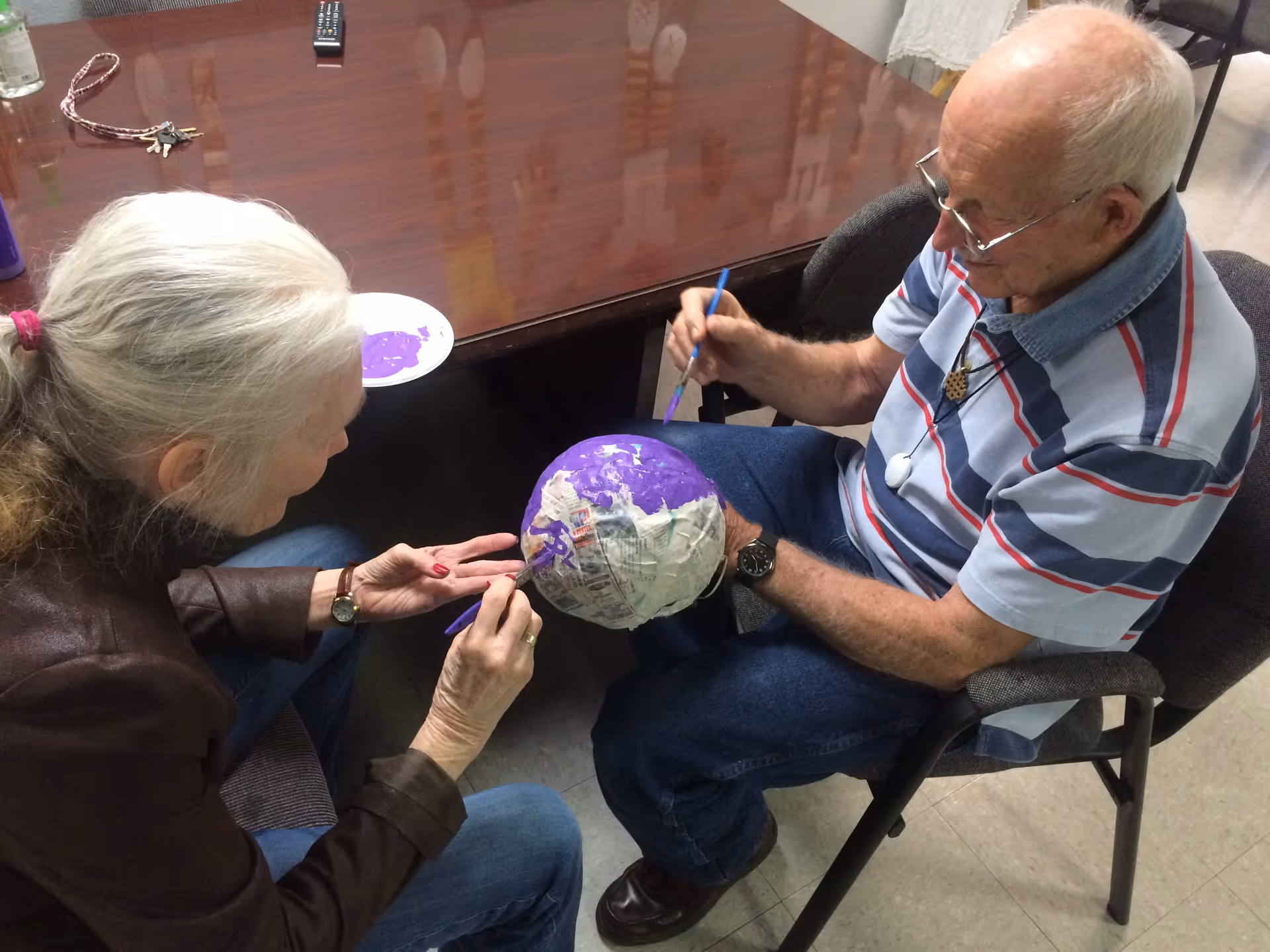 Two elderly individuals sitting at a table engaged in a craft activity, painting a papier-mâché globe with purple paint. The woman on the left is holding the globe while the man on the right is painting it with a brush. Various items including keys and a remote control are on the table.