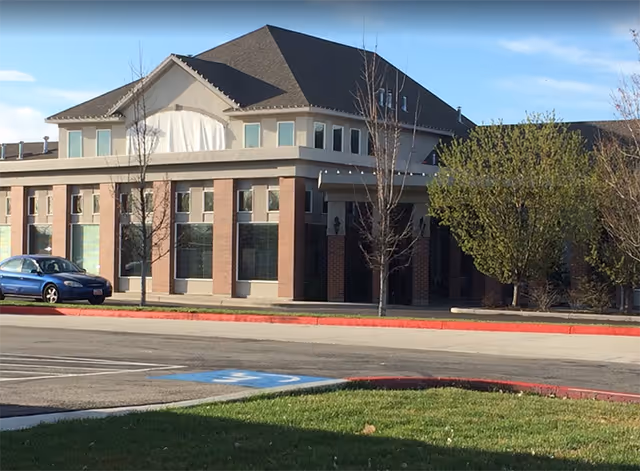 Front exterior of a two-story assisted living building with brick columns, large windows, trees, and a parked blue car in the lot.