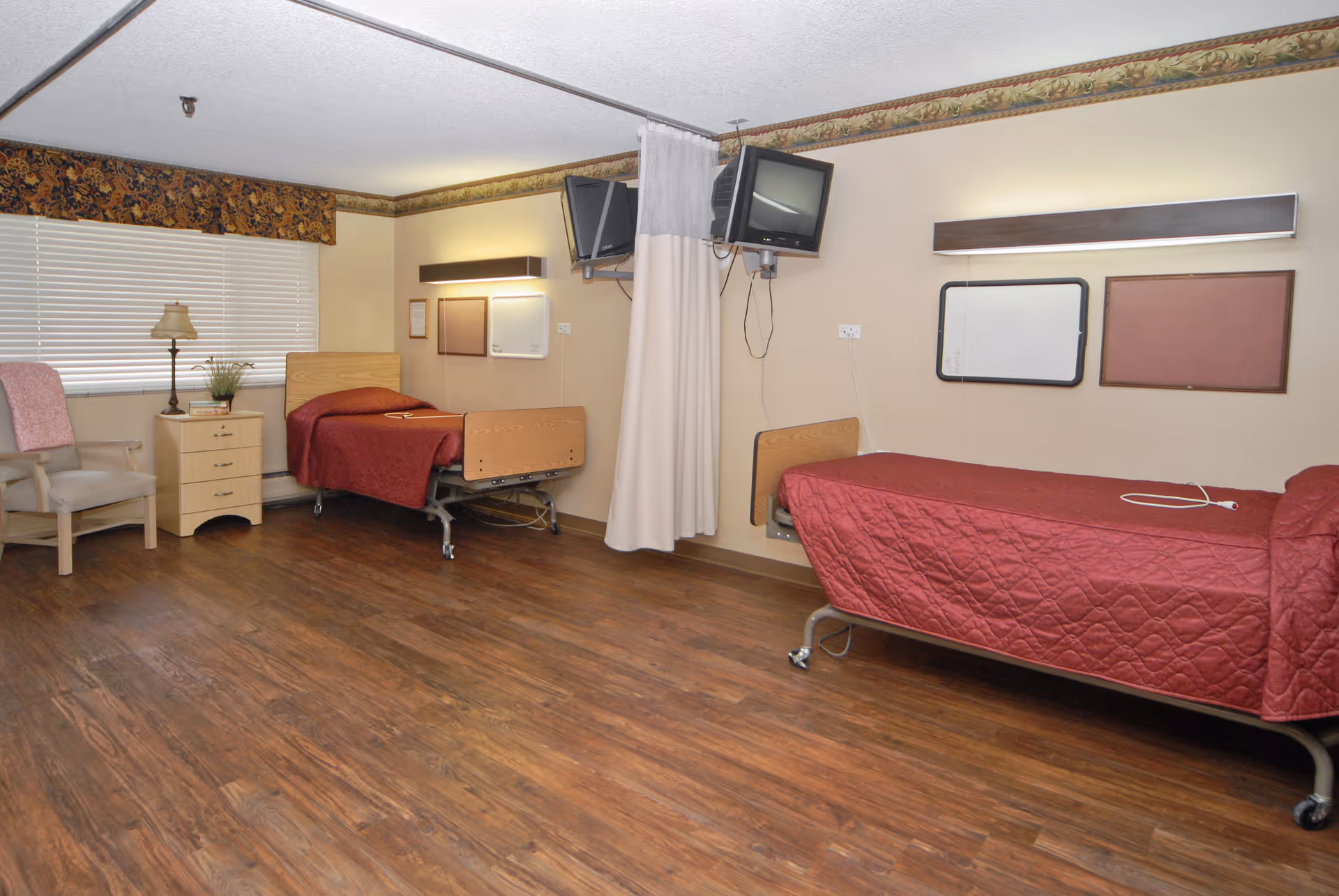 A room with two hospital-style beds covered with red quilts, a wooden nightstand with a lamp and plant, a beige armchair with a pink cushion, two wall-mounted televisions, a privacy curtain between the beds, and hardwood flooring.
