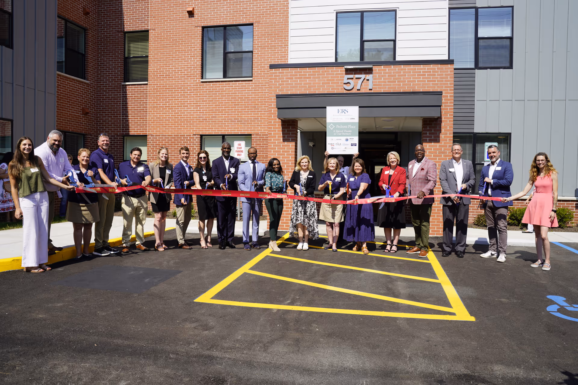 A diverse group of people stands in front of a brick senior-living building holding a red ribbon during a ribbon-cutting ceremony.