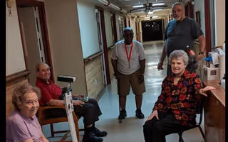 Four elderly individuals gathered in a hallway of a senior living facility. Two are seated on chairs near the walls, one is standing in the middle of the hallway, and another is standing near a counter. The hallway is well-lit with ceiling lights and has handrails along the walls.