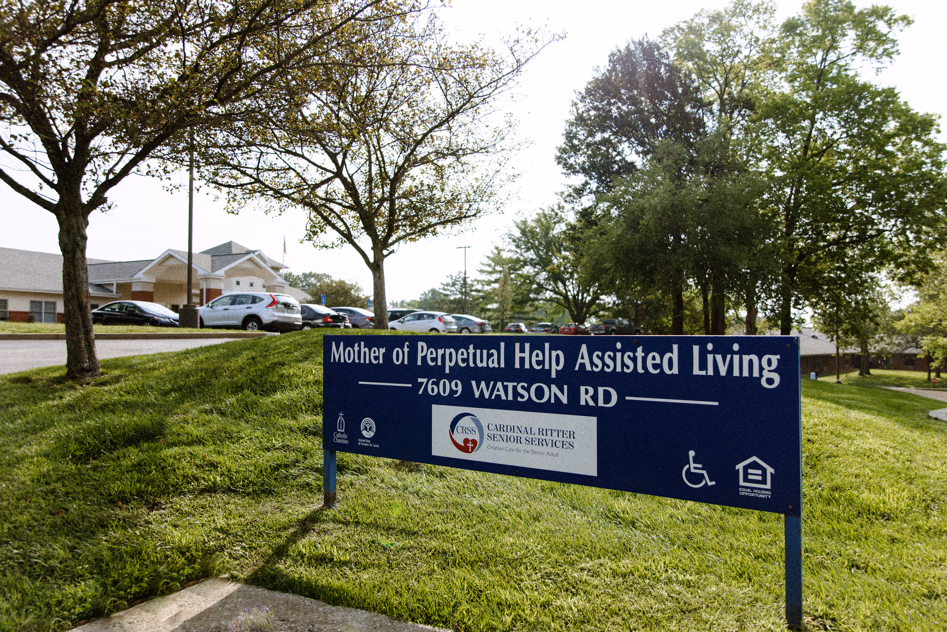 A blue lawn sign for Mother of Perpetual Help Assisted Living at 7609 Watson Rd in front of the facility building, parked cars and trees.