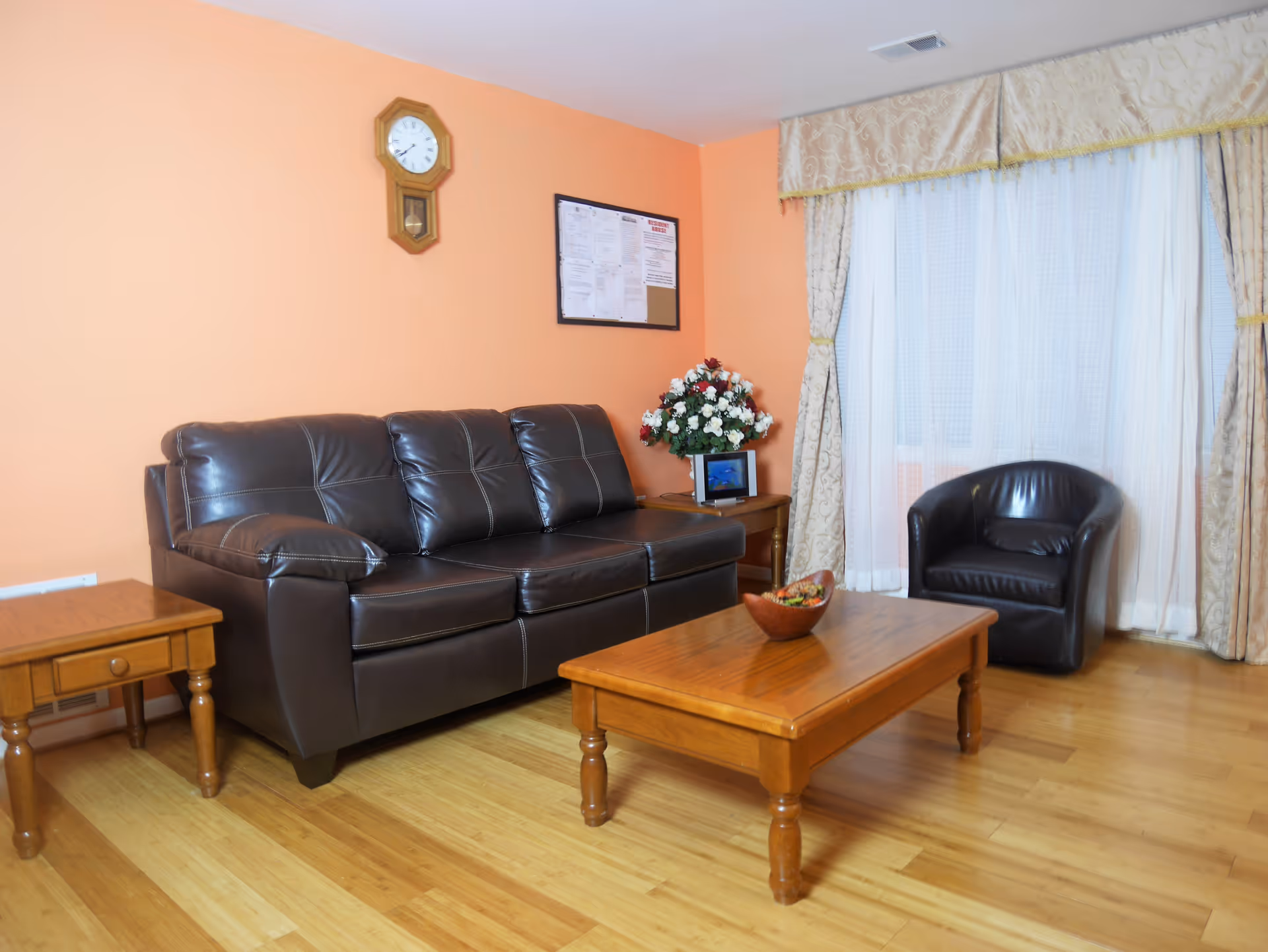 A cozy living room with a dark brown leather sofa and matching armchair. There is a wooden coffee table with a decorative bowl on it, and a wooden side table with a vase of flowers and a small framed picture. The walls are painted peach, with a wooden clock and a bulletin board hanging on the wall. Large windows with sheer white curtains and patterned drapes allow natural light into the room. The floor is light wood.