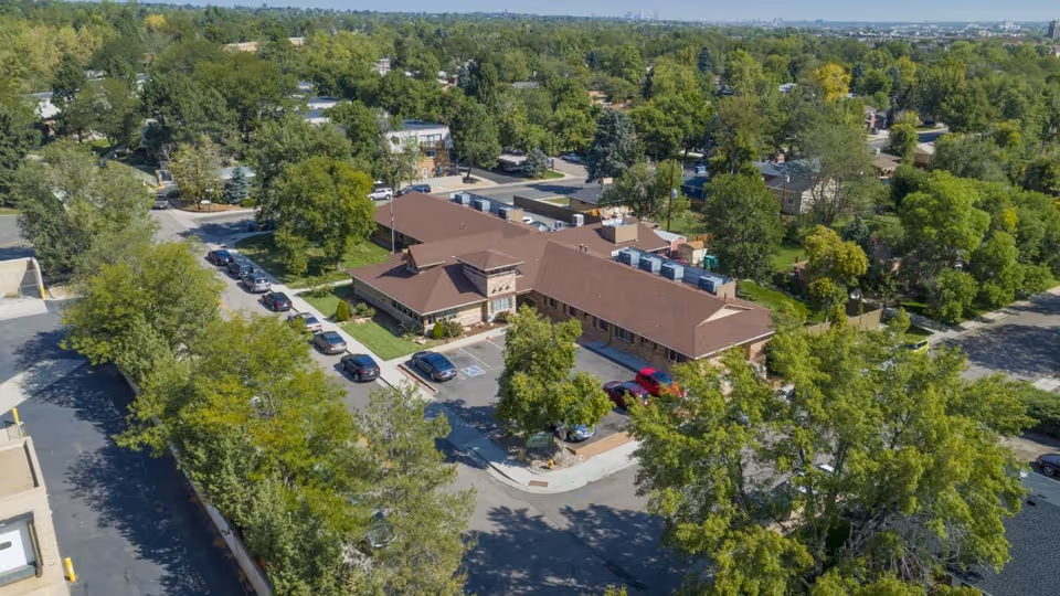 Aerial view of a single-story building with a brown roof surrounded by trees and parked cars in a suburban neighborhood. The building is situated at a street corner with greenery and residential houses in the background.