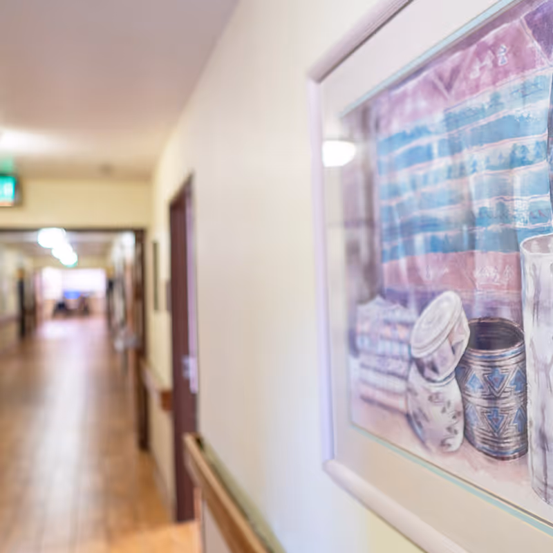 A long indoor hallway with wooden flooring and beige walls, featuring a framed painting of pottery and textiles on the right wall. The hallway has several doors and handrails along the walls, with bright ceiling lights and an exit sign visible in the distance.
