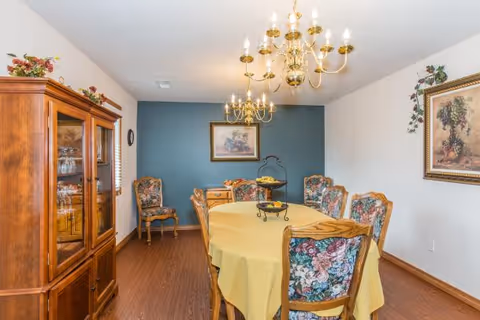A dining room with a wooden table covered by a yellow tablecloth surrounded by six floral upholstered chairs. Two brass chandeliers hang from the ceiling above the table. A wooden china cabinet with glass doors is on the left side, and framed artwork hangs on the walls. The floor is wood, and the far wall is painted dark teal.
