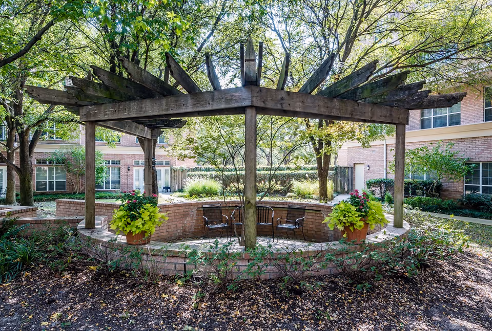 Outdoor seating area with a circular brick wall and a wooden pergola overhead. There are four metal chairs inside the circular area and two large flower pots with green and pink plants on the brick wall. The area is surrounded by trees and shrubs, with a building visible in the background.