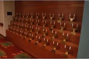 Wood-paneled interior wall with stepped shelves holding multiple identical brass holders arranged in rows.