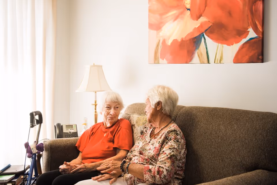 Two elderly women sitting and talking on a brown couch in a well-lit living room with a lamp and a large floral painting on the wall behind them.