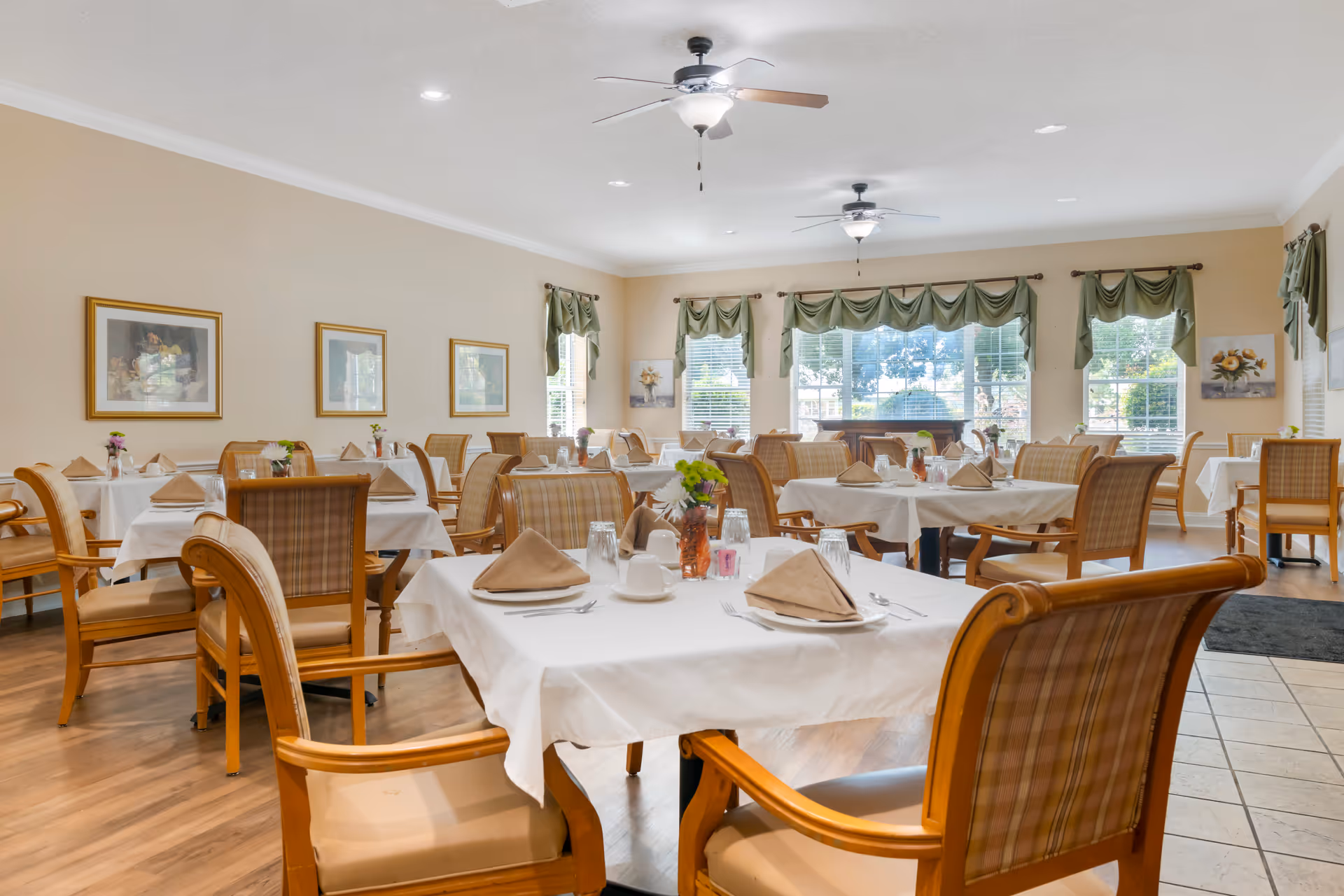 A bright dining room with multiple tables covered in white tablecloths, each set with beige napkins, cups, glasses, and silverware. Wooden chairs with plaid upholstery surround the tables. The room has large windows with green valances, ceiling fans, framed artwork on the walls, and a wooden floor.