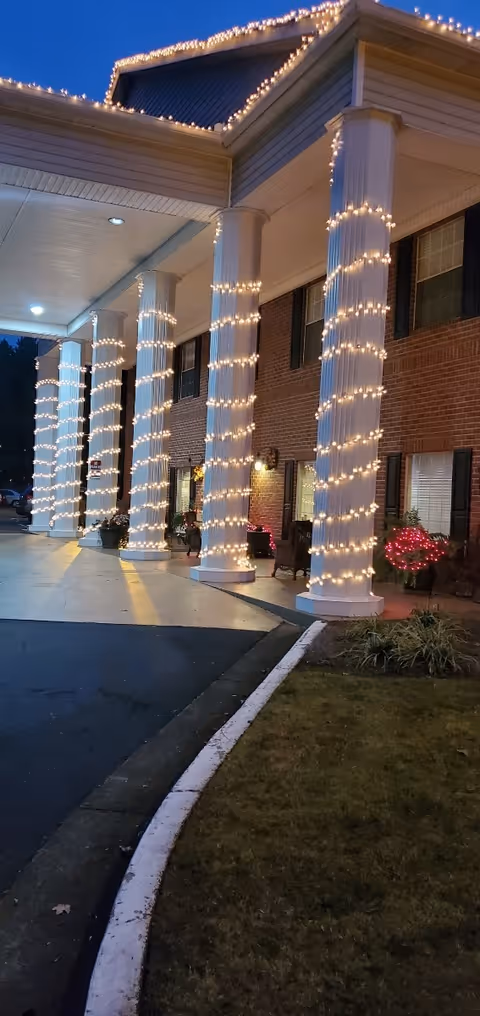 Entrance of a building at dusk with tall white columns wrapped in white string lights. The building has a brick exterior with windows and black shutters. There are small bushes decorated with red lights near the entrance and a covered driveway with a curved curb.