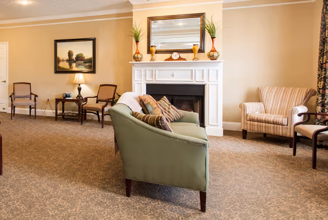 A cozy living room area in a senior living facility featuring a green upholstered sofa with striped pillows, a white fireplace with decorative vases and a clock on the mantel, a large mirror above the fireplace, a side table with a lamp, two wooden chairs, a striped armchair, and a framed landscape painting on the wall.