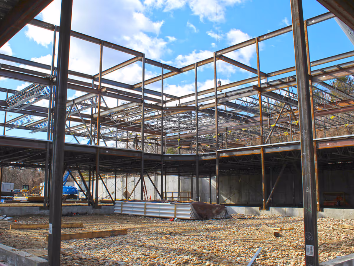 Interior view of a building under construction showing exposed steel framework, trusses, and a gravel floor.