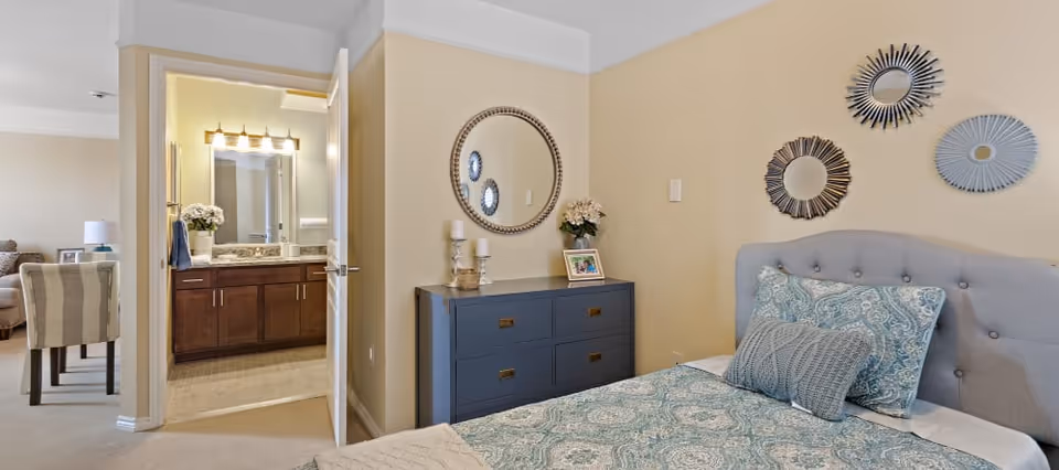 A bedroom with a bed featuring a blue and white patterned comforter and pillows. On the right wall above the bed are three decorative round mirrors. Next to the bed is a dark blue dresser with candles, a framed photo, and a flower vase. An open door reveals a bathroom with a vanity, sink, and mirror with lights. In the background, part of a living room with a striped chair and a lamp is visible.
