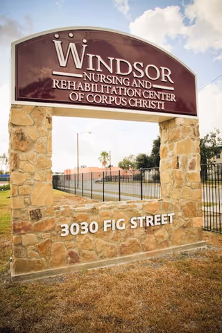 Stone sign for Windsor Nursing and Rehabilitation Center of Corpus Christi with address 3030 Fig Street, set outdoors with a partly cloudy sky in the background.