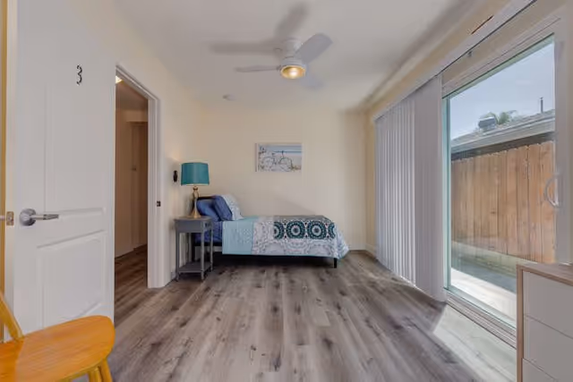 A bright bedroom with a single bed against the wall, decorated with blue and teal bedding. There is a small gray nightstand with a blue lamp next to the bed. A framed picture hangs above the bed. The room has light-colored walls, wood-look flooring, a ceiling fan with a light, and a sliding glass door with vertical blinds leading to an outdoor area. A wooden chair is partially visible in the foreground.