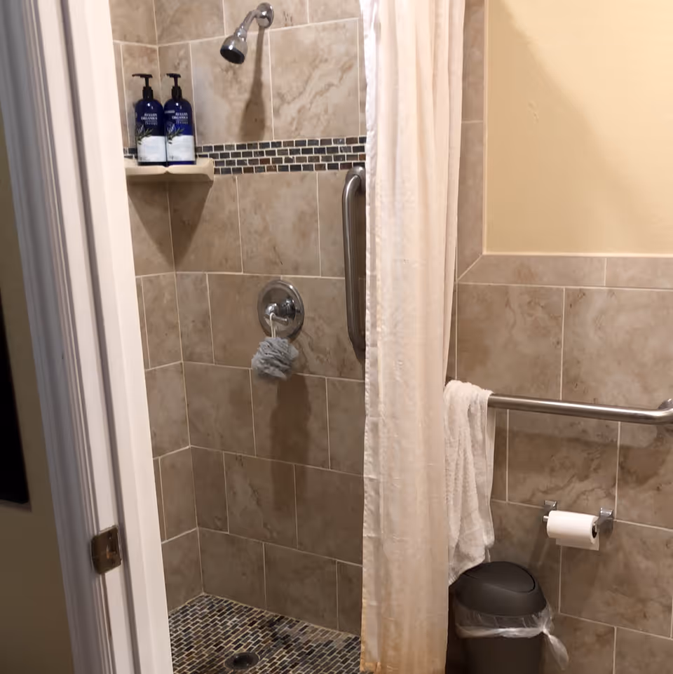 A tiled shower area with a silver showerhead, a grab bar, and a white shower curtain partially drawn. There are two blue bottles of soap or shampoo on a small corner shelf. A gray bath sponge hangs from the shower control. Outside the shower, a towel hangs on a grab bar above a trash can with a plastic liner, and a toilet paper holder is mounted on the wall.