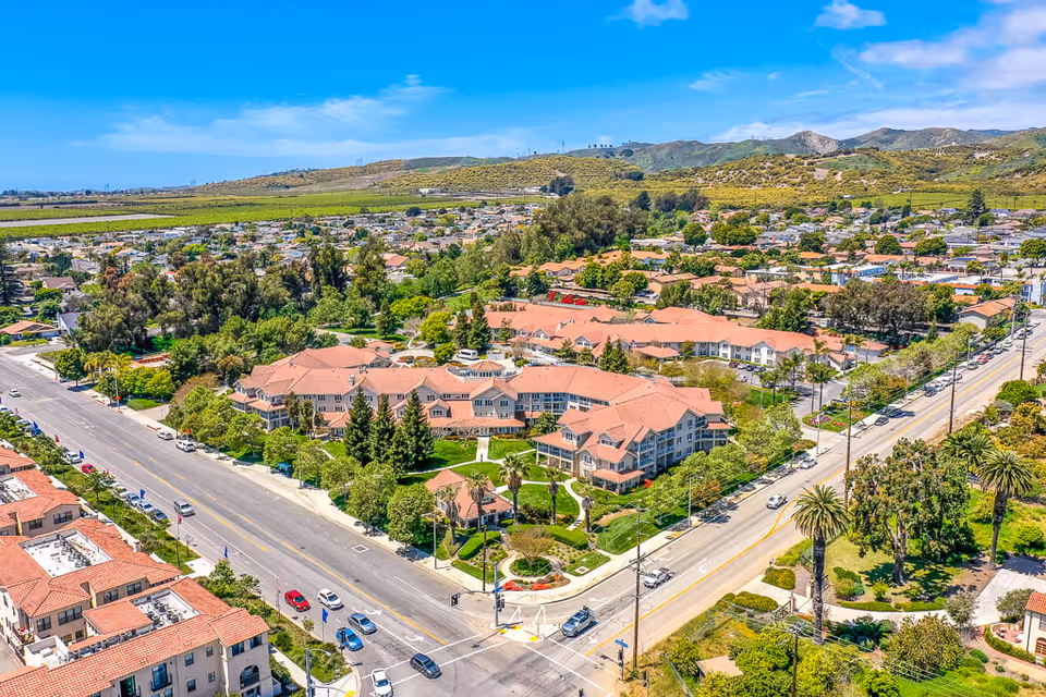 Aerial view of The Palms At Bonaventure Assisted Living & Memory Care facility surrounded by trees and greenery, with a large building complex featuring red-tiled roofs. The facility is located at a busy intersection with cars on the roads and residential neighborhoods and hills in the background under a blue sky with some clouds.