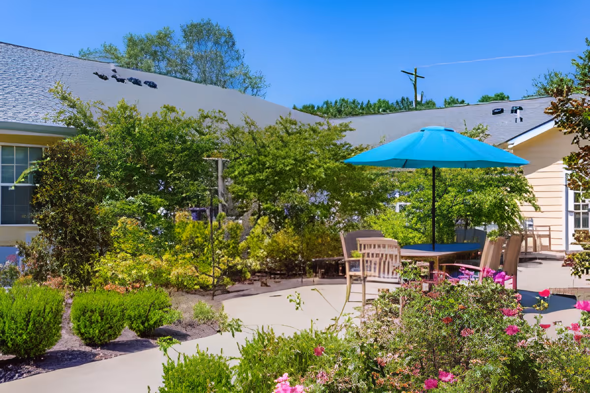 Outdoor patio area at Charter Senior Living of Hermitage with a round table, several chairs, and a large blue umbrella surrounded by lush green bushes and flowering plants, with the building and clear blue sky in the background.