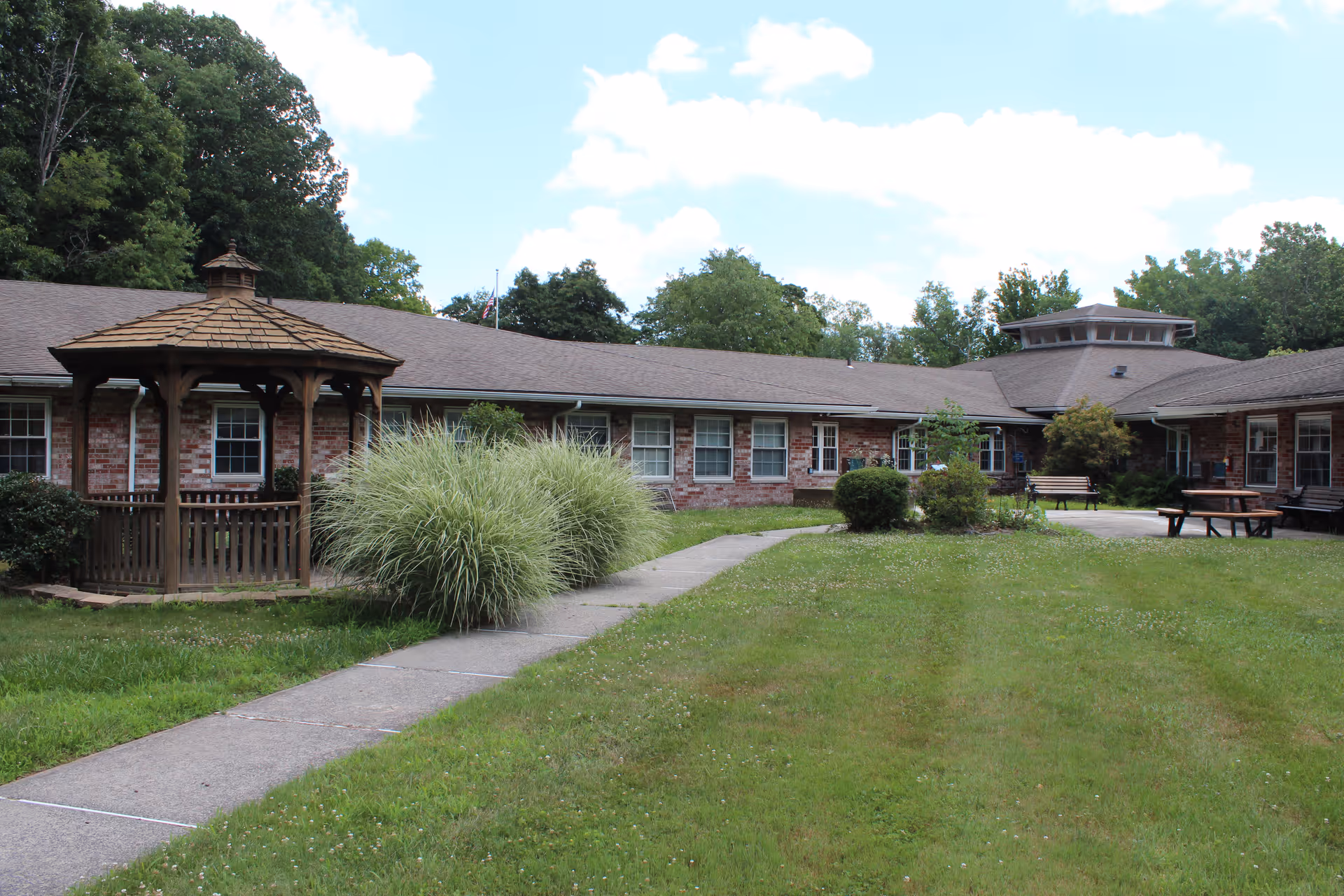 Outdoor courtyard area of a senior living facility with a wooden gazebo on the left, a concrete walkway, green grass, bushes, and trees surrounding a single-story brick building with multiple windows and benches.